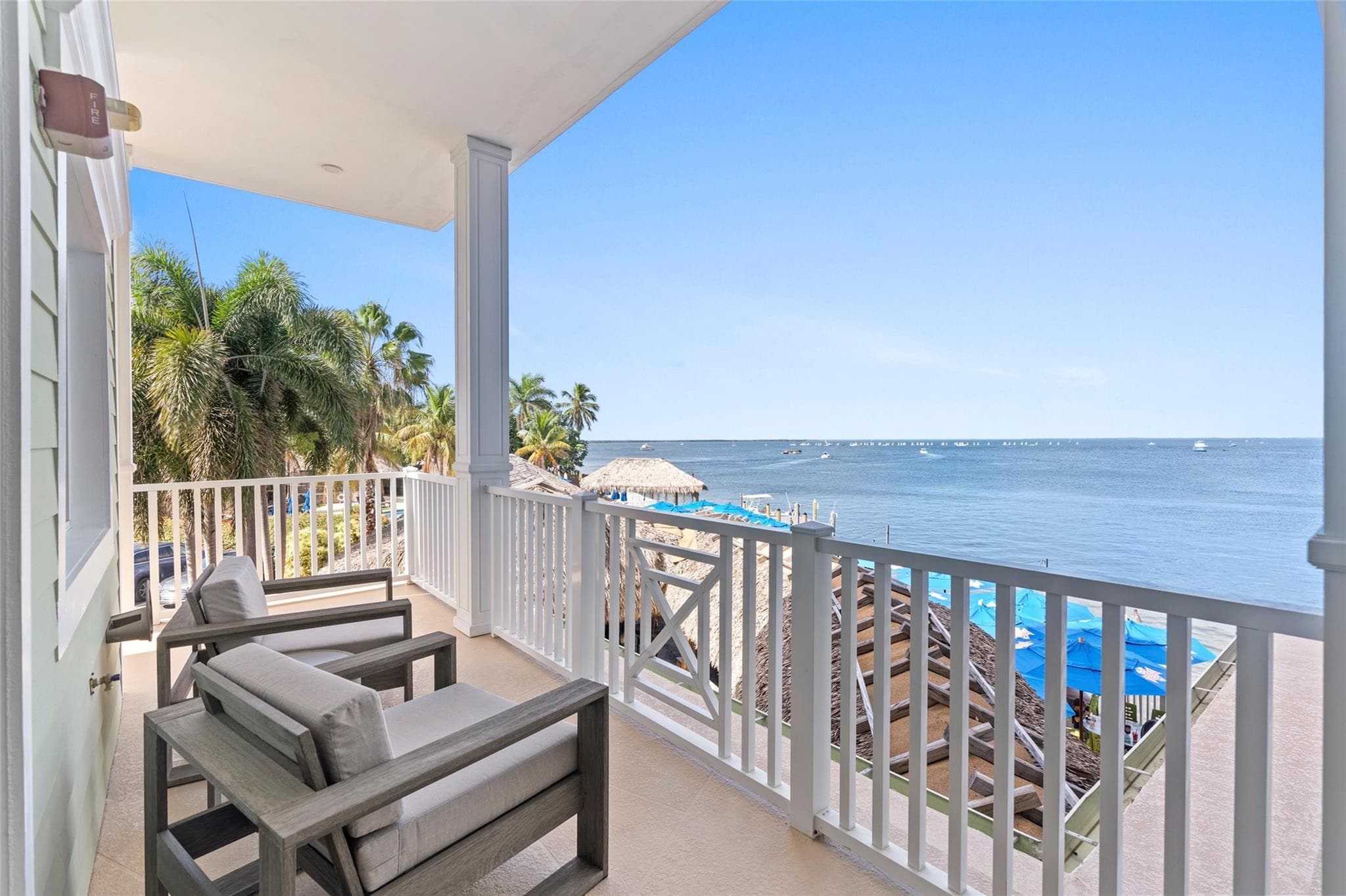 Private balcony with two gray cushioned armchairs looking down over thatched palapa roofs, blue beach umbrellas, and boats scattered across the bay.