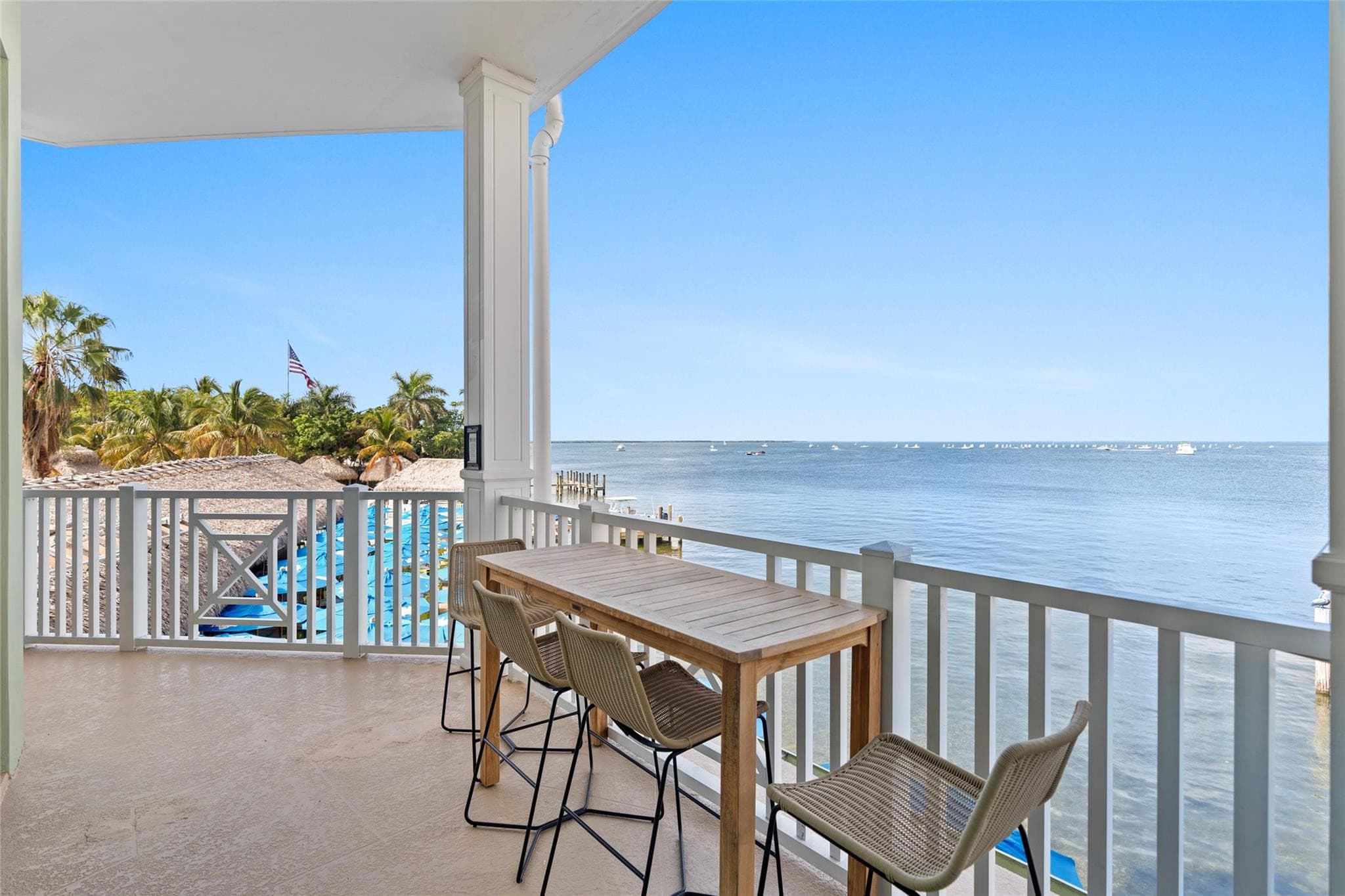 Corner balcony with a teak pub table and woven stools overlooking blue umbrellas, thatched roofs, a wooden dock, and an American flag along the bayfront.