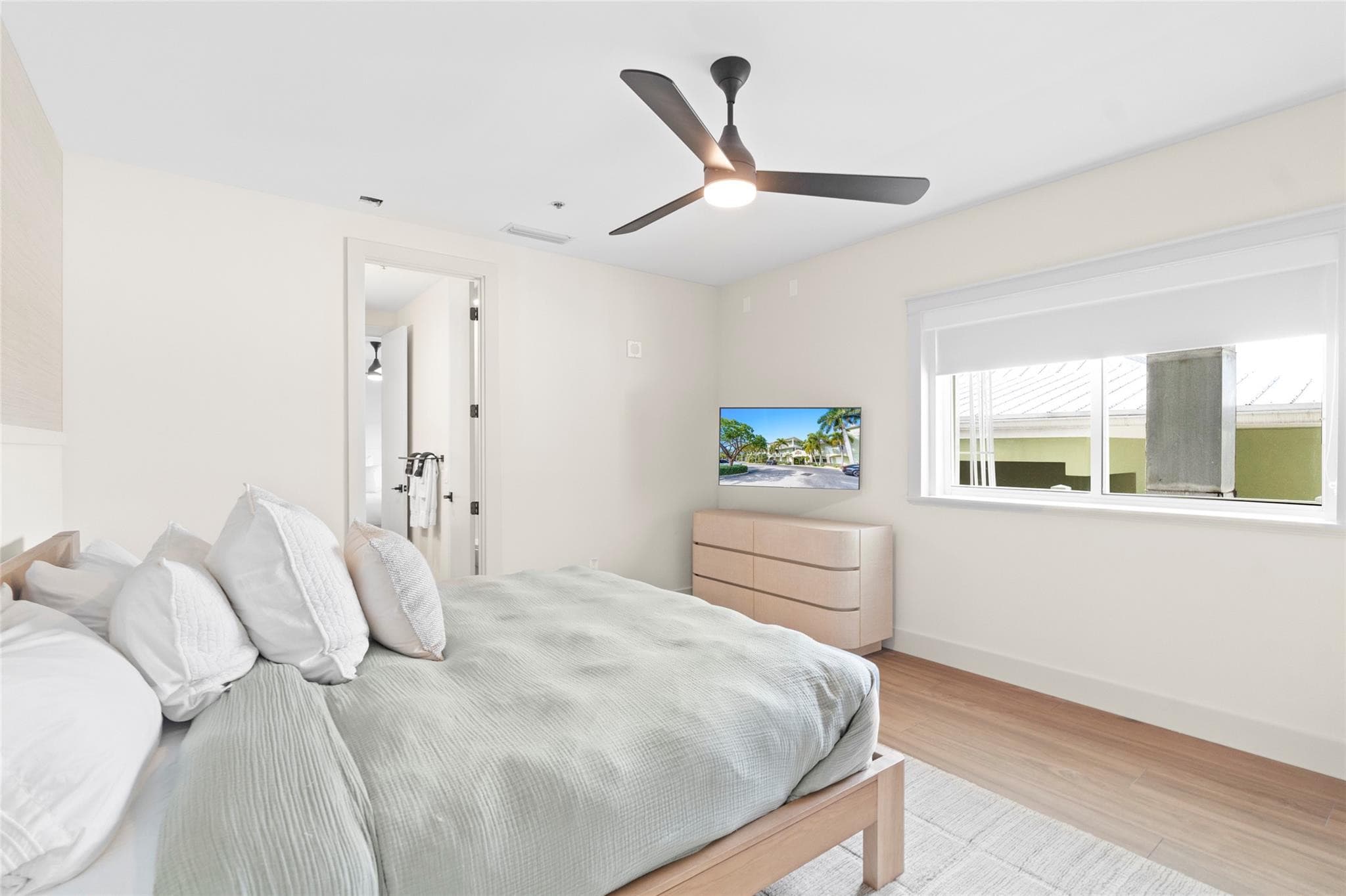 Guest bedroom with sage bedding and stacked white pillows, a light-wood dresser below a wall-mounted TV, ceiling fan, and a wide window with roller shade.