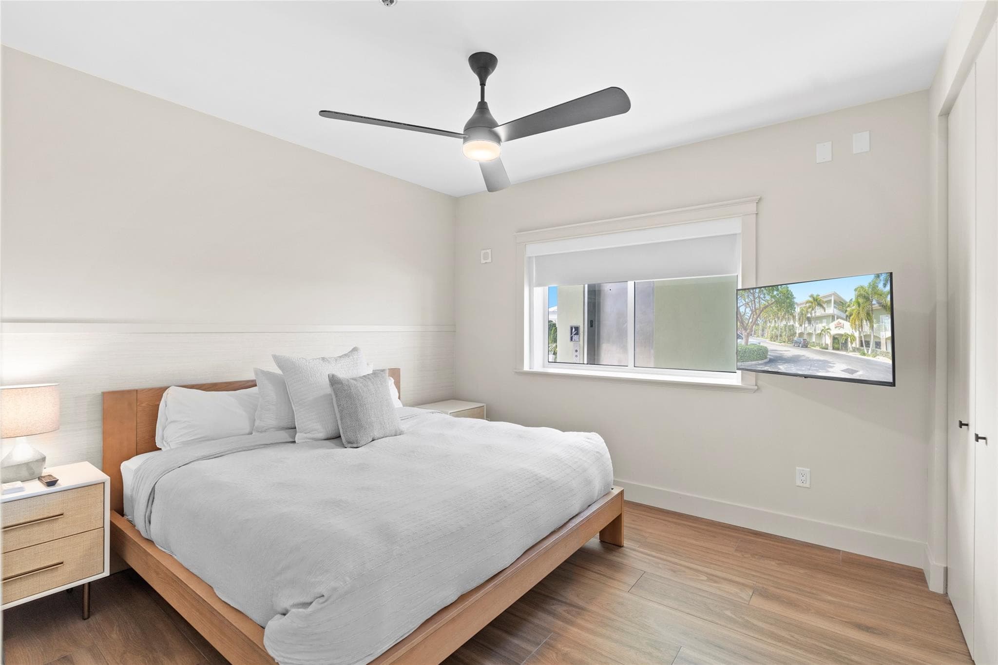 Guest bedroom with a light-wood king bed, crisp white bedding, gray accent pillow, woven-front nightstands, and a wall-mounted TV beside the window.