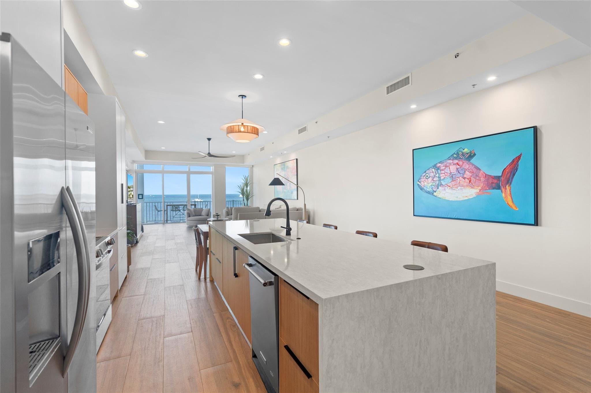 Galley-style kitchen with a long gray quartz island, stainless refrigerator and dishwasher, and a vivid mosaic-style fish painting on the wall, looking toward the water-view living room.