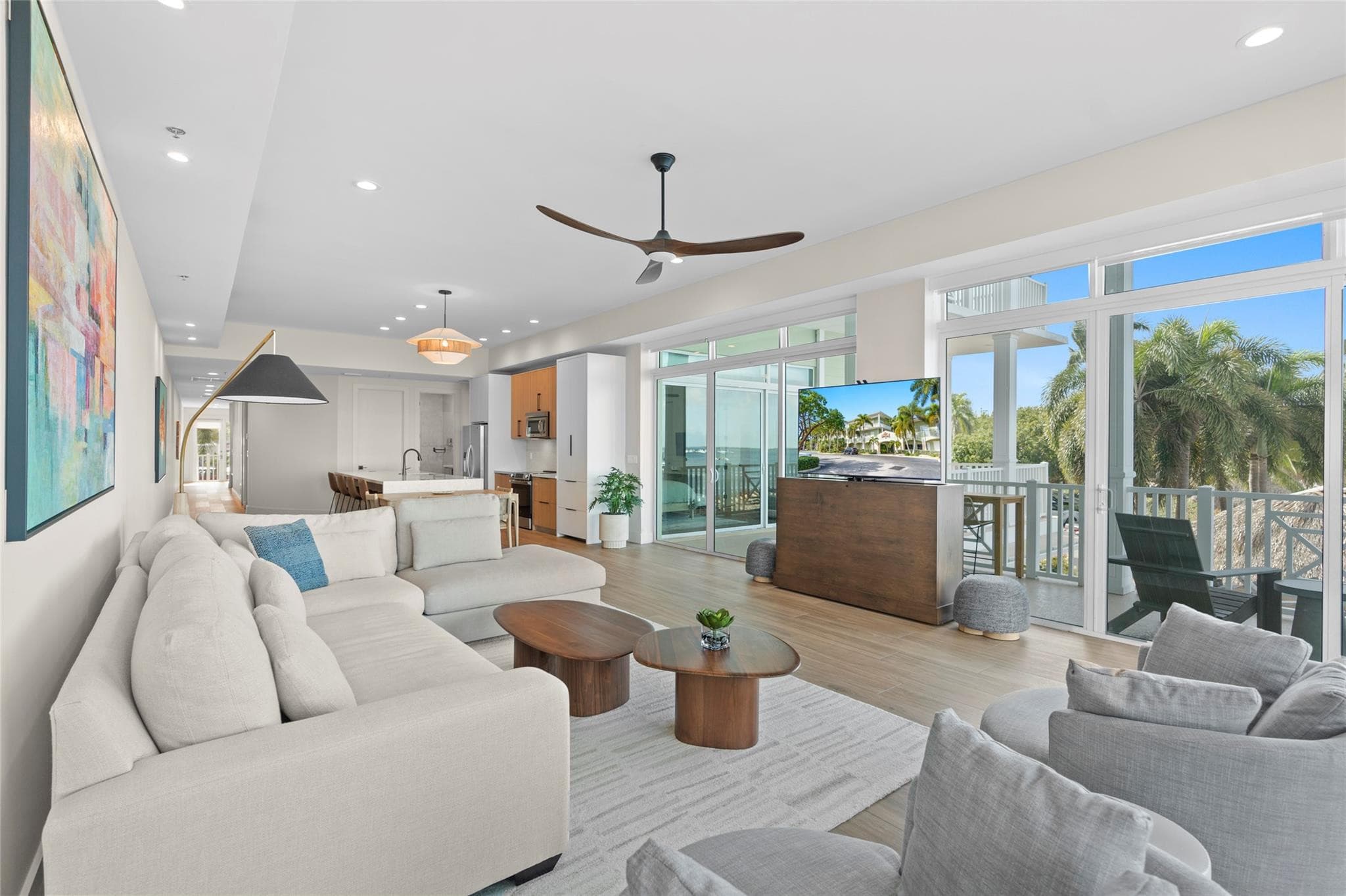 Bright living room with a cream sectional, walnut coffee tables, colorful abstract art, a TV lift cabinet, and sliding glass doors to a covered balcony framed by palms.