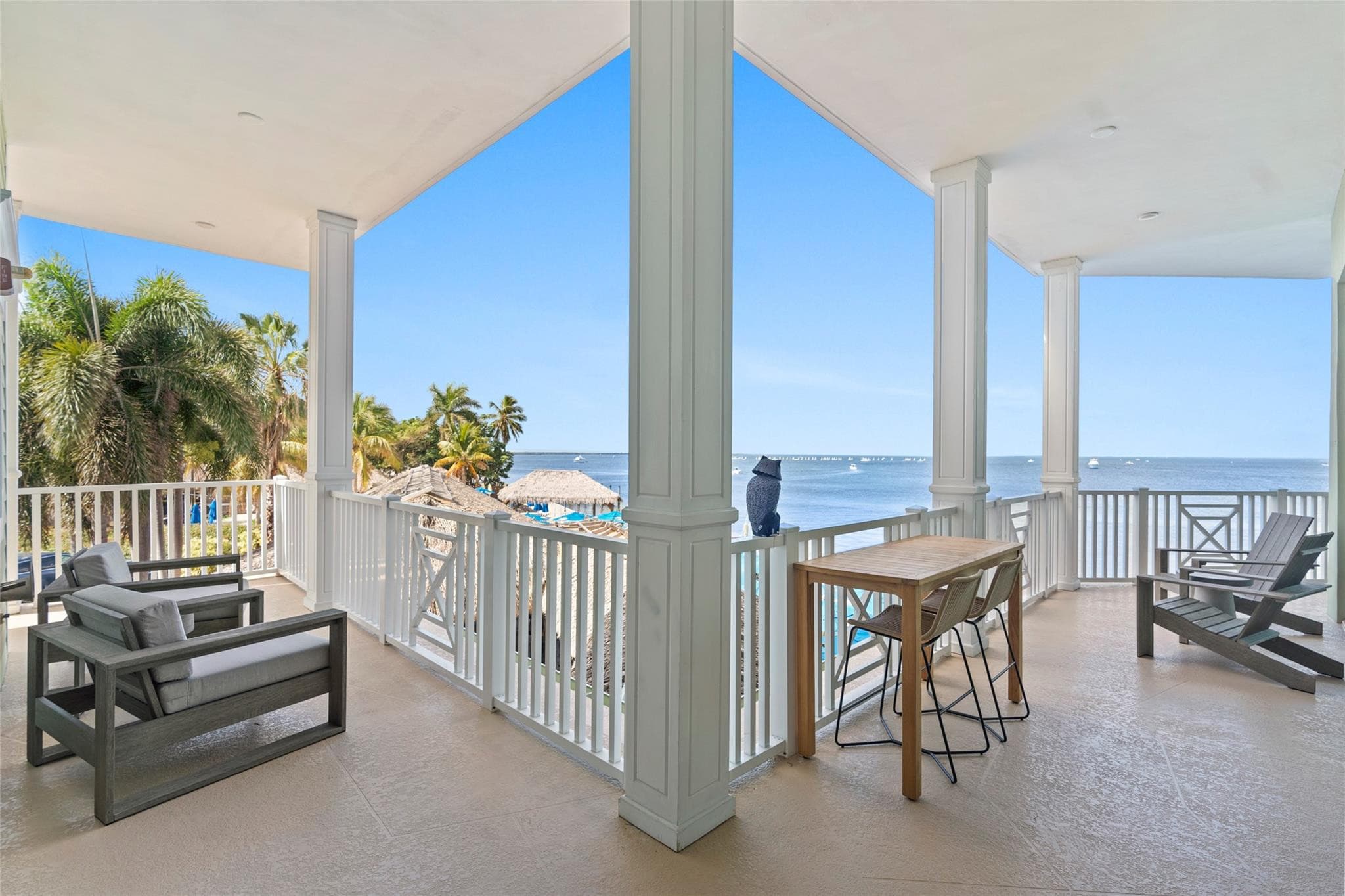 Expansive wraparound covered balcony with white columns, gray cushioned lounge furniture, teak bar table, and Adirondack chairs overlooking thatched tiki roofs and open water.